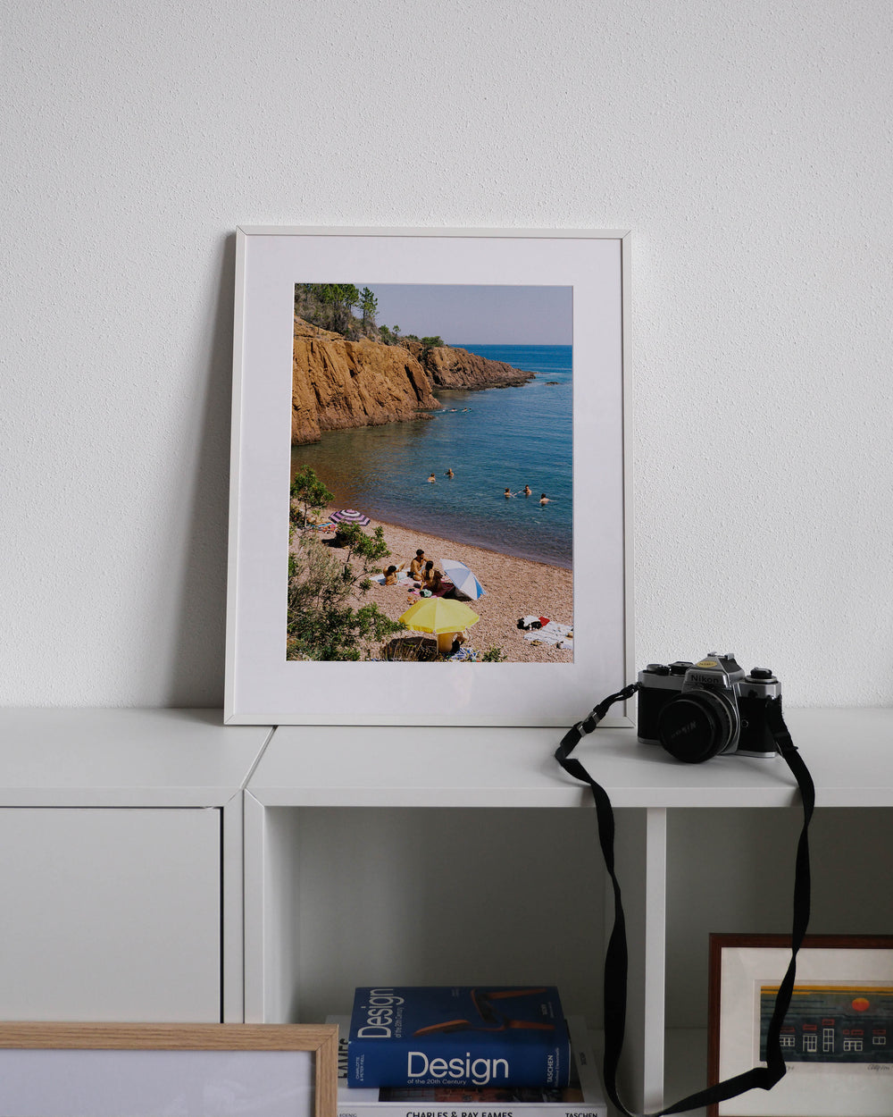 Framed analog photo print on Fujifilm paper featuring Cap Roux, with parasols shading sunbathers on warm stones and others cooling off in the Mediterranean, set against the striking contrast of red rocks and the blue sea, displayed on a modern white cabinet with a minimalist interior setting.