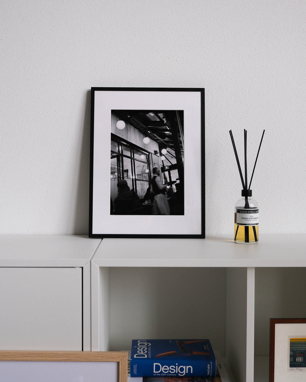 Framed black-and-white analog photo print on Fujifilm paper featuring the iconic Café du Flore in Saint-Germain-des-Prés, with a busy waiter serving guests amidst the café's classic Parisian ambiance. Displayed on a modern white cabinet with a minimalist interior setting.