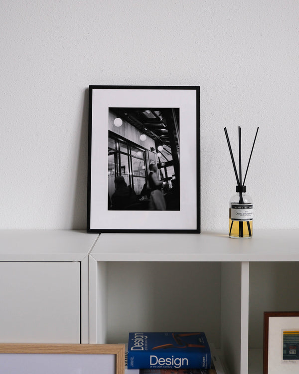Framed black-and-white analog photo print on Fujifilm paper featuring the iconic Café du Flore in Saint-Germain-des-Prés, with a busy waiter serving guests amidst the café's classic Parisian ambiance. Displayed on a modern white cabinet with a minimalist interior setting.