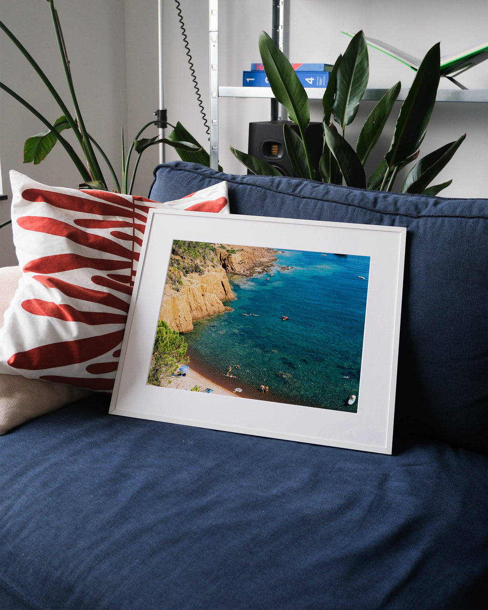 Framed analog photo print on Fujifilm paper featuring the vibrant contrast of the blue Mediterranean waters and red rocks of Cap Roux, with people enjoying the clear sea on the Côte d'Azur, displayed on a blue couch with a minimalist interior setting.