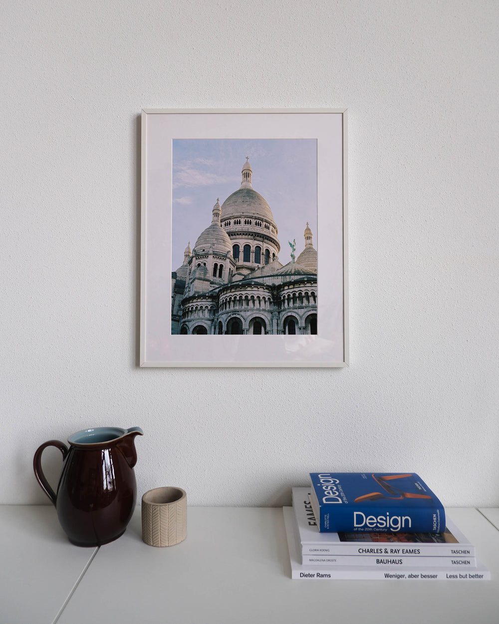 Framed analog photo print on Fujifilm paper featuring the towers of Sacré-Cœur in winter, bathed in sunlight with a clear blue sky above, displayed on a modern white cabinet with a minimalist interior setting.