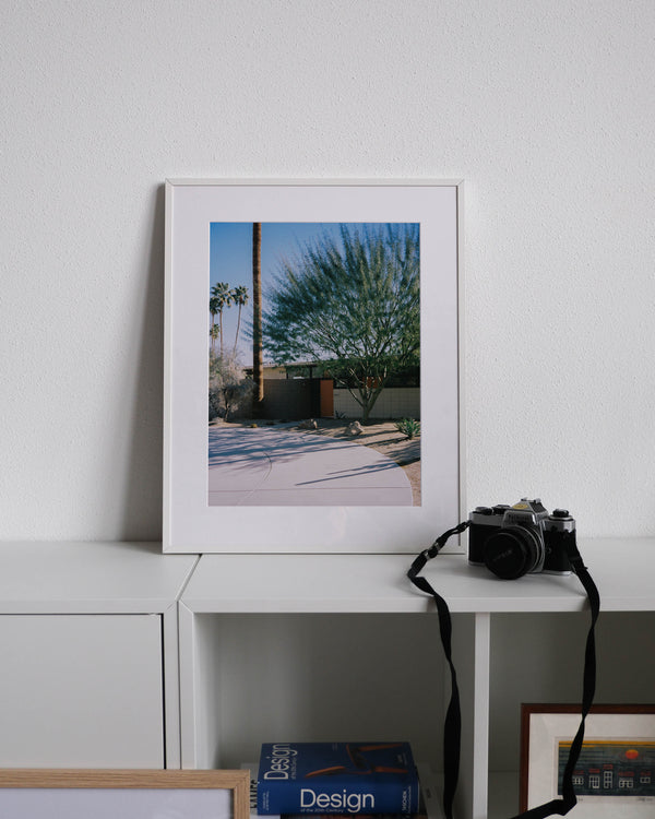 Framed analog photo print on Fujifilm paper featuring featuring a tree in Palm Springs, positioned in front of a midcentury modern home, displayed on a modern white cabinet with a minimalist interior setting.