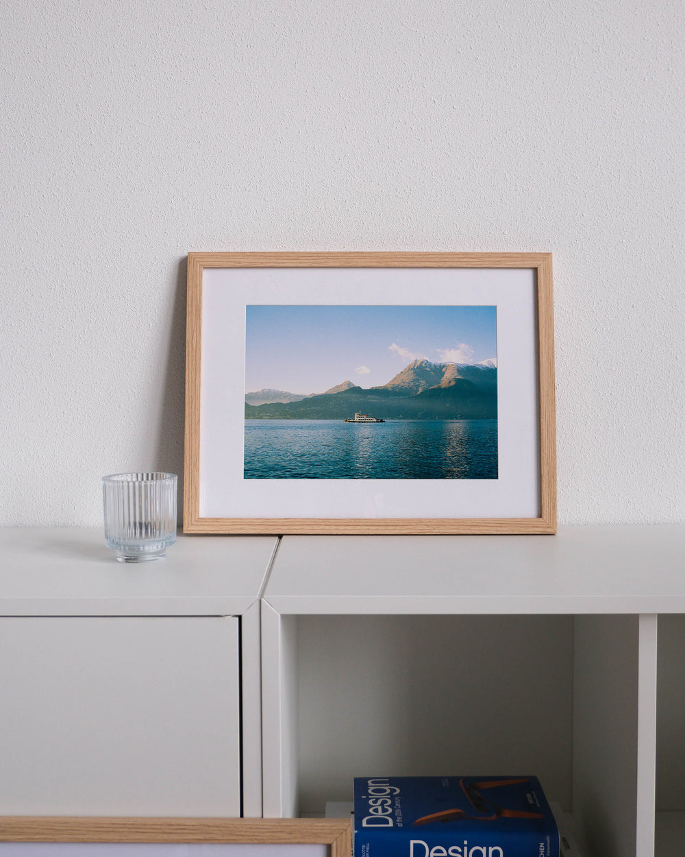 Framed analog photo print on Fujifilm paper featuring a ferry crossing Lake Como in winter in Bellano, with mountain peak Monte Bregagno in the background, displayed on a modern white cabinet with a minimalist interior setting.