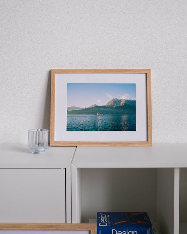 Framed analog photo print on Fujifilm paper featuring a ferry crossing Lake Como in winter in Bellano, with mountain peak Monte Bregagno in the background, displayed on a modern white cabinet with a minimalist interior setting.