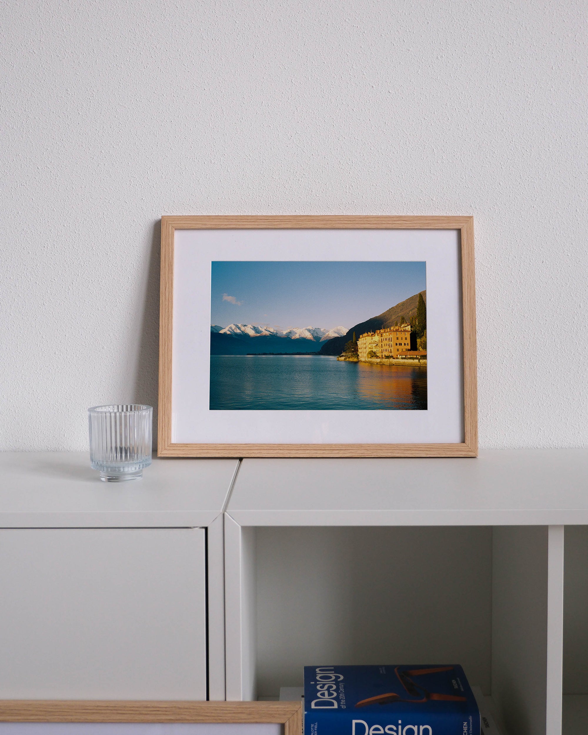Framed analog photo print on Fujifilm paper featuring houses along Lake Como in Bellano during golden hour, with snow-capped peaks, displayed on a modern white cabinet with a minimalist interior setting.
