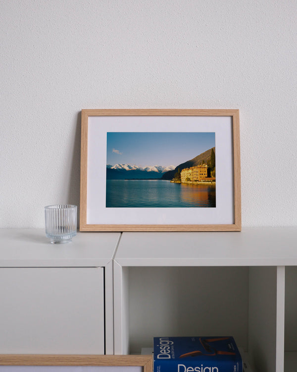 Framed analog photo print on Fujifilm paper featuring houses along Lake Como in Bellano during golden hour, with snow-capped peaks, displayed on a modern white cabinet with a minimalist interior setting.