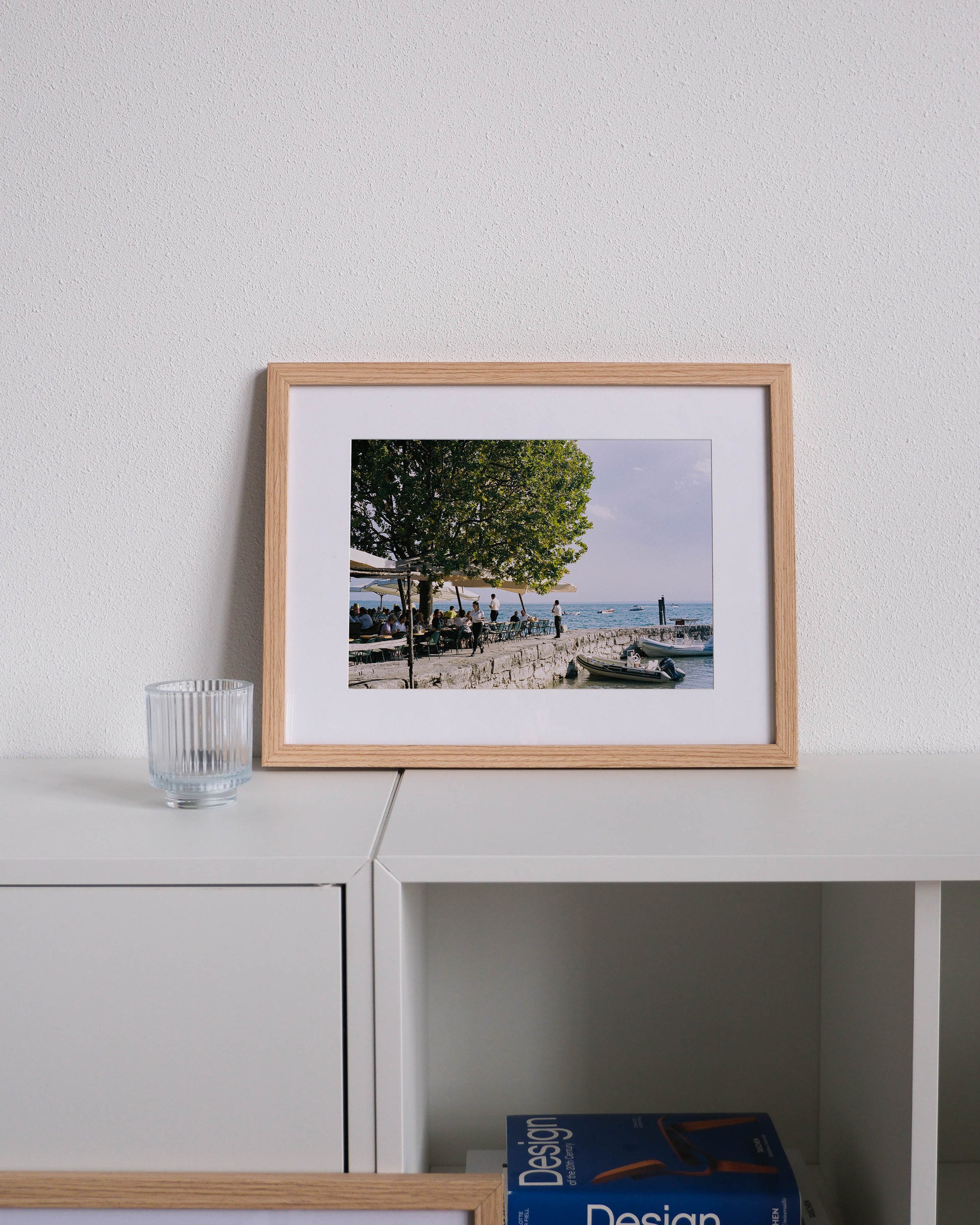Framed analog photo print on Fujifilm paper featuring a serene lakeside terrace on Lake Garda, with waiters serving guests under a tree, soft sunlight filtering through the leaves, and the tranquil waters in the background. Displayed on a modern white cabinet with a minimalist interior setting.