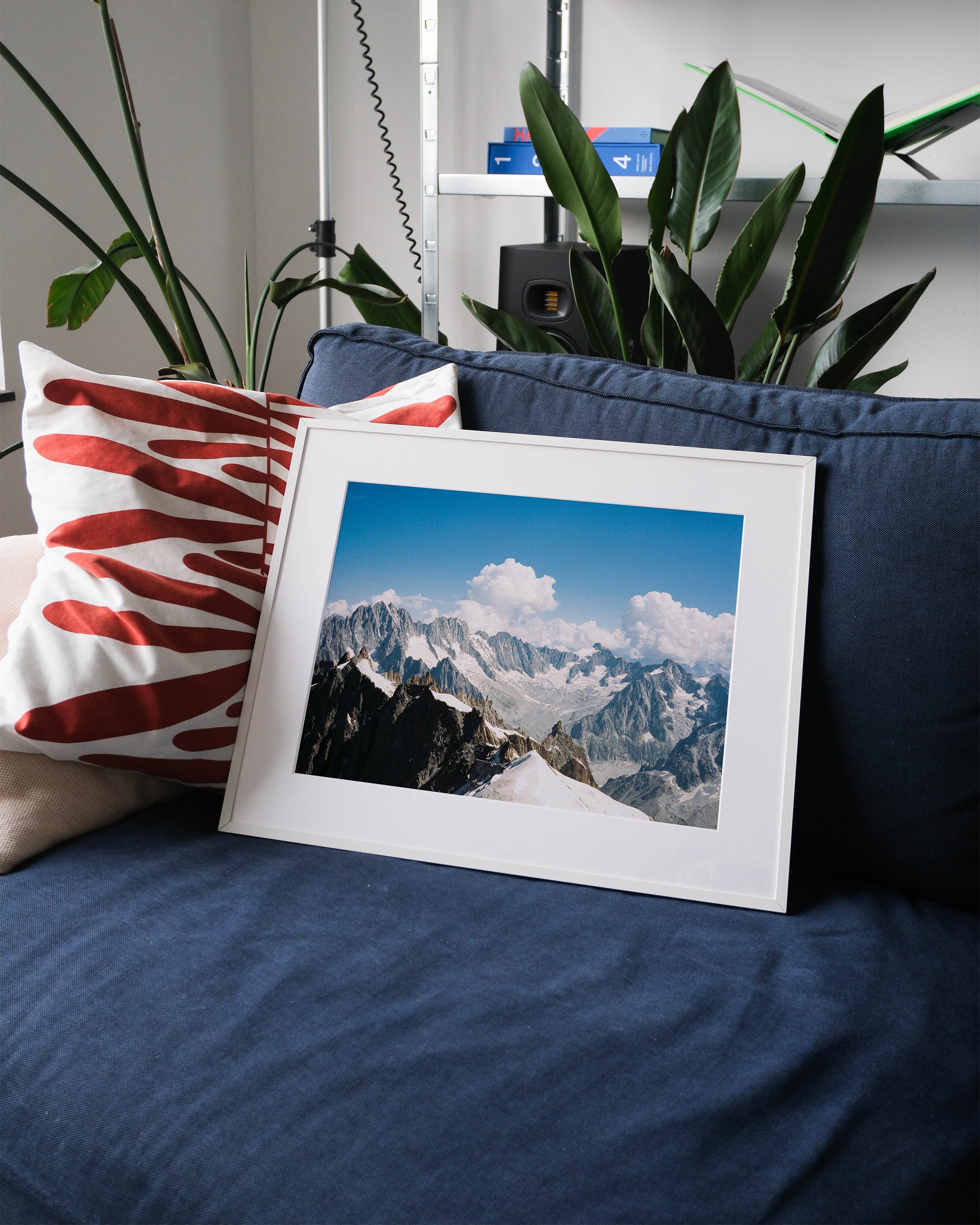 Framed analog photo print on Fujifilm paper featuring the Mont Blanc massif, captured from the Aiguille du Midi at 3842 meters. The steep peaks rise against a blue sky, with swirling clouds adding contrast to the rugged landscape. Captured in Chamonix. Displayed on a blue couch with a minimalist interior setting.