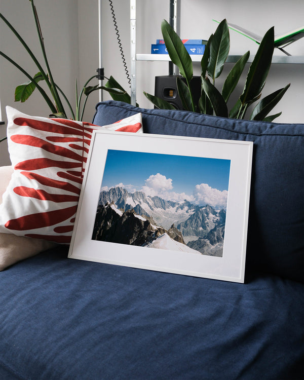 Framed analog photo print on Fujifilm paper featuring the Mont Blanc massif, captured from the Aiguille du Midi at 3842 meters. The steep peaks rise against a blue sky, with swirling clouds adding contrast to the rugged landscape. Captured in Chamonix. Displayed on a blue couch with a minimalist interior setting.