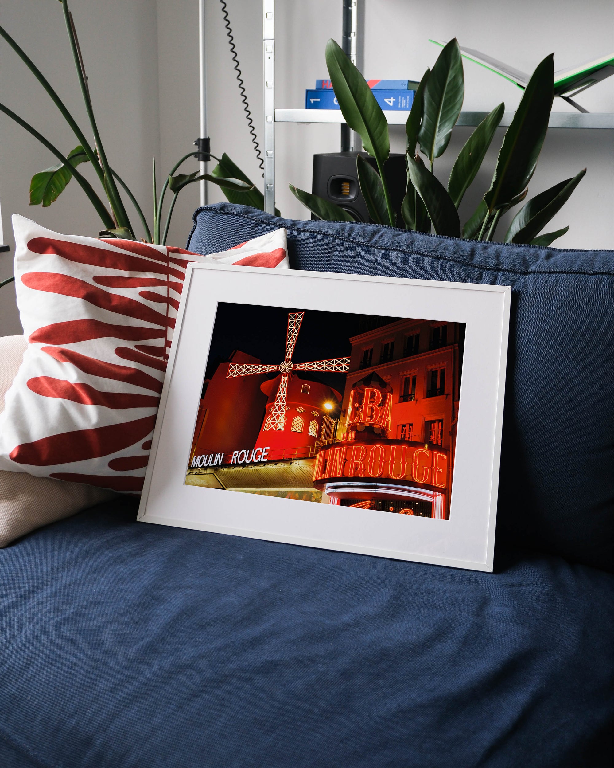 Framed analog photo print on Fujifilm paper featuring  the iconic Moulin Rouge at night, with its bright red lights glowing against the Parisian night sky, displayed on a blue couch with a minimalist interior setting.