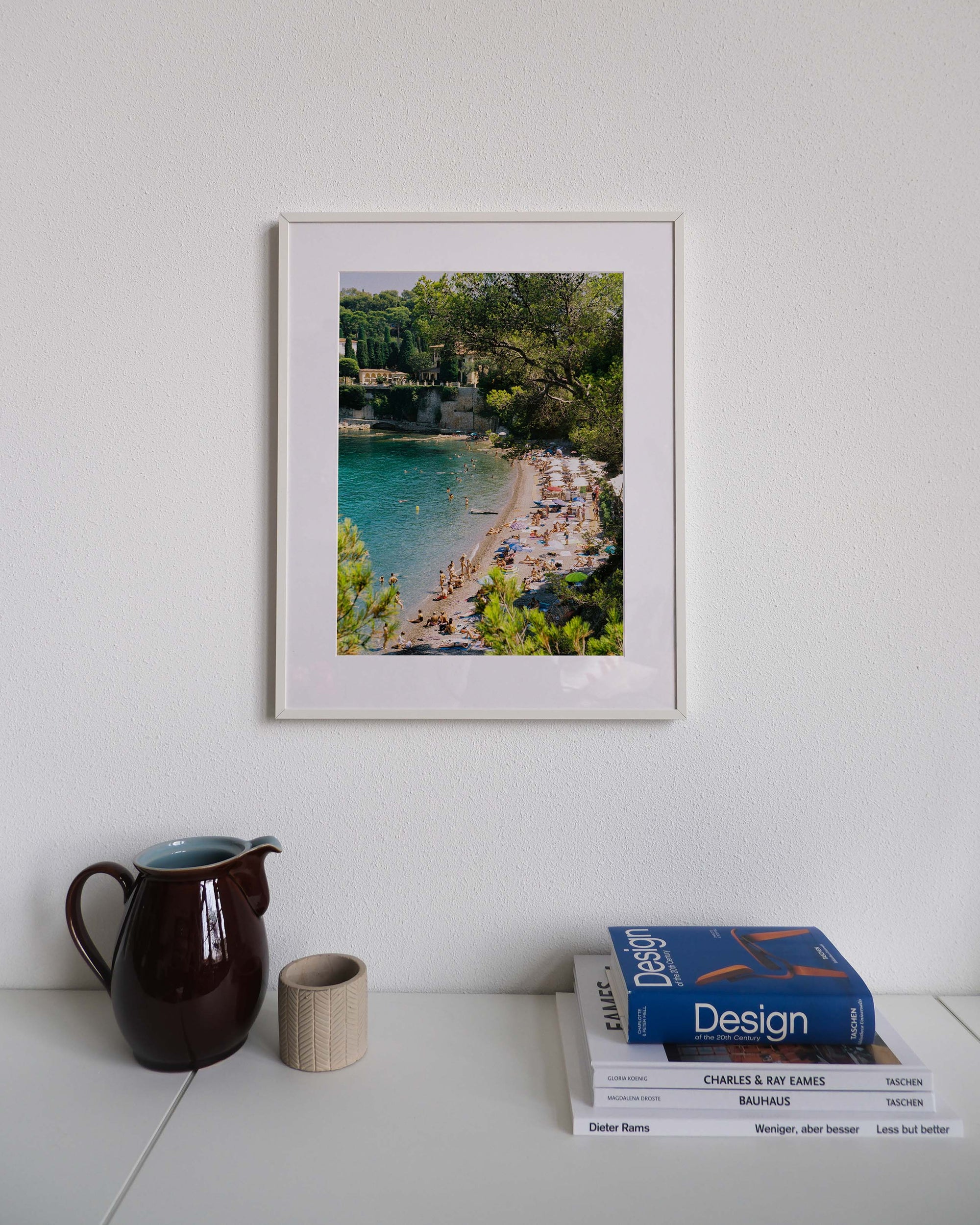 Framed analog photo print on Fujifilm paper featuring Paloma Beach in Saint-Jean-Cap-Ferrat, with sunbathers on the sands, the Mediterranean Sea, and luxurious villas along the coastline, displayed on a modern white cabinet with a minimalist interior setting.