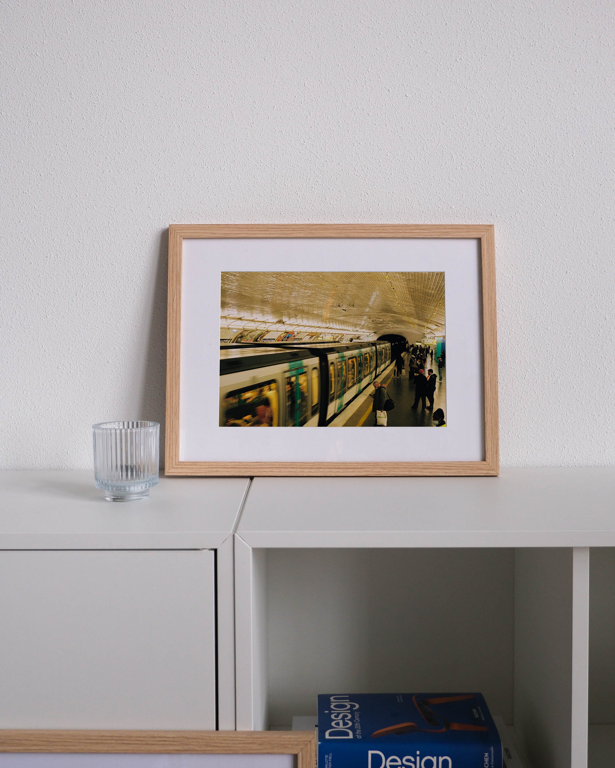 Framed analog photo print on Fujifilm paper featuring a scene inside the Paris metro, with a man checking his phone while waiting for the subway. Displayed on a modern white cabinet with a minimalist interior setting.