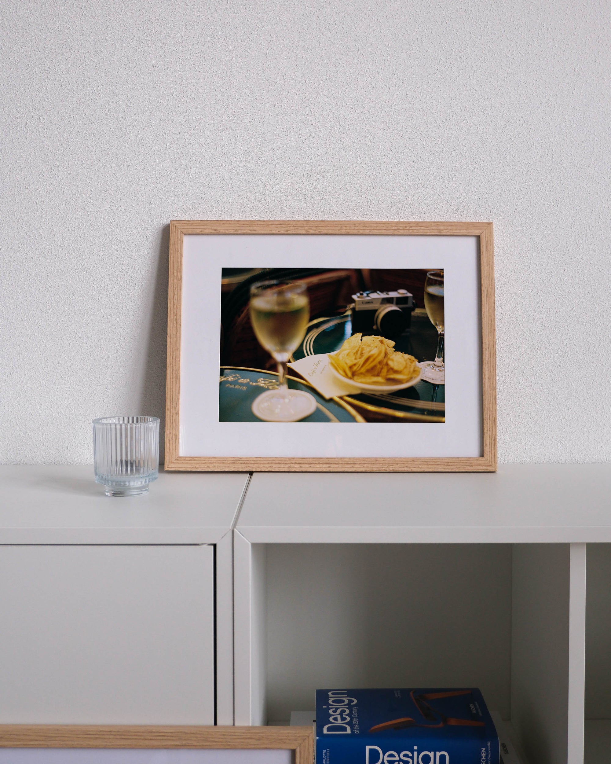 Framed analog photo print on Fujifilm paper  a close-up of an engraved table at Café du Flore, with a glass of white wine, a bowl of chips, and an analog camera, displayed on a modern white cabinet with a minimalist interior setting.