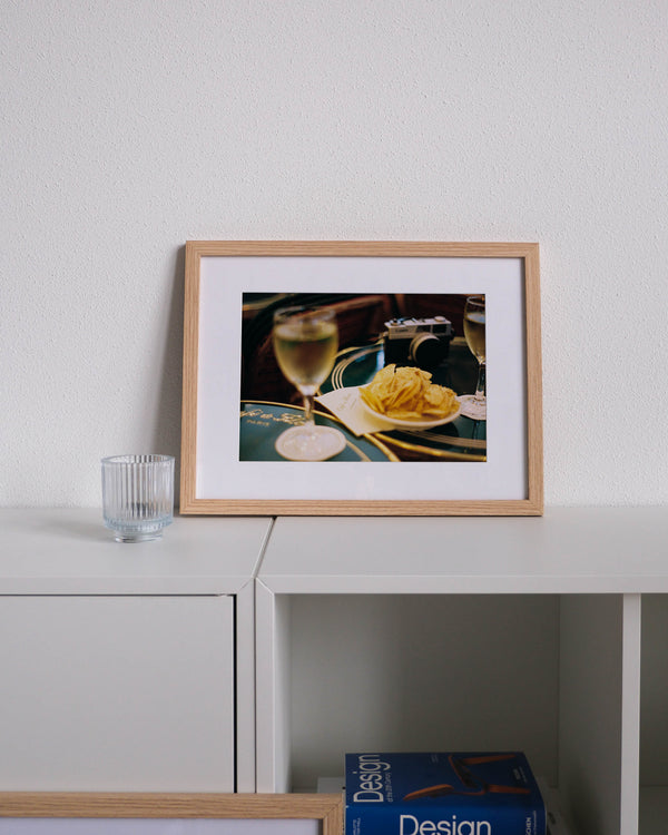 Framed analog photo print on Fujifilm paper  a close-up of an engraved table at Café du Flore, with a glass of white wine, a bowl of chips, and an analog camera, displayed on a modern white cabinet with a minimalist interior setting.