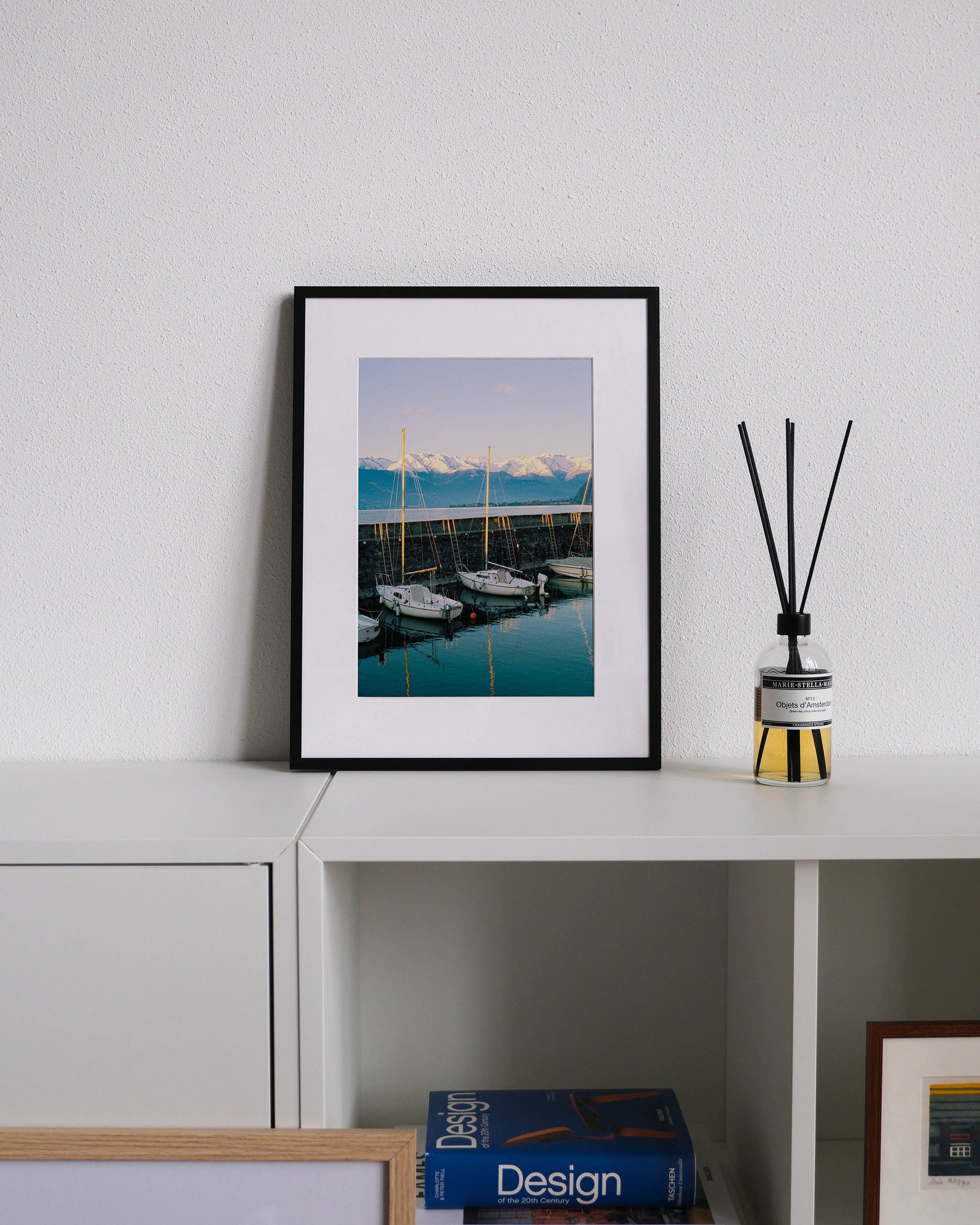 Framed analog photo print on Fujifilm paper featuring sailboats resting on the still water in Bellano Marina on a winter day, with mountains in the background and a warm sunset glow, displayed on a modern white cabinet with a minimalist interior setting.