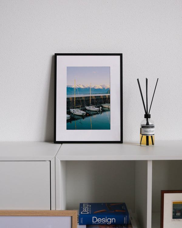 Framed analog photo print on Fujifilm paper featuring sailboats resting on the still water in Bellano Marina on a winter day, with mountains in the background and a warm sunset glow, displayed on a modern white cabinet with a minimalist interior setting.