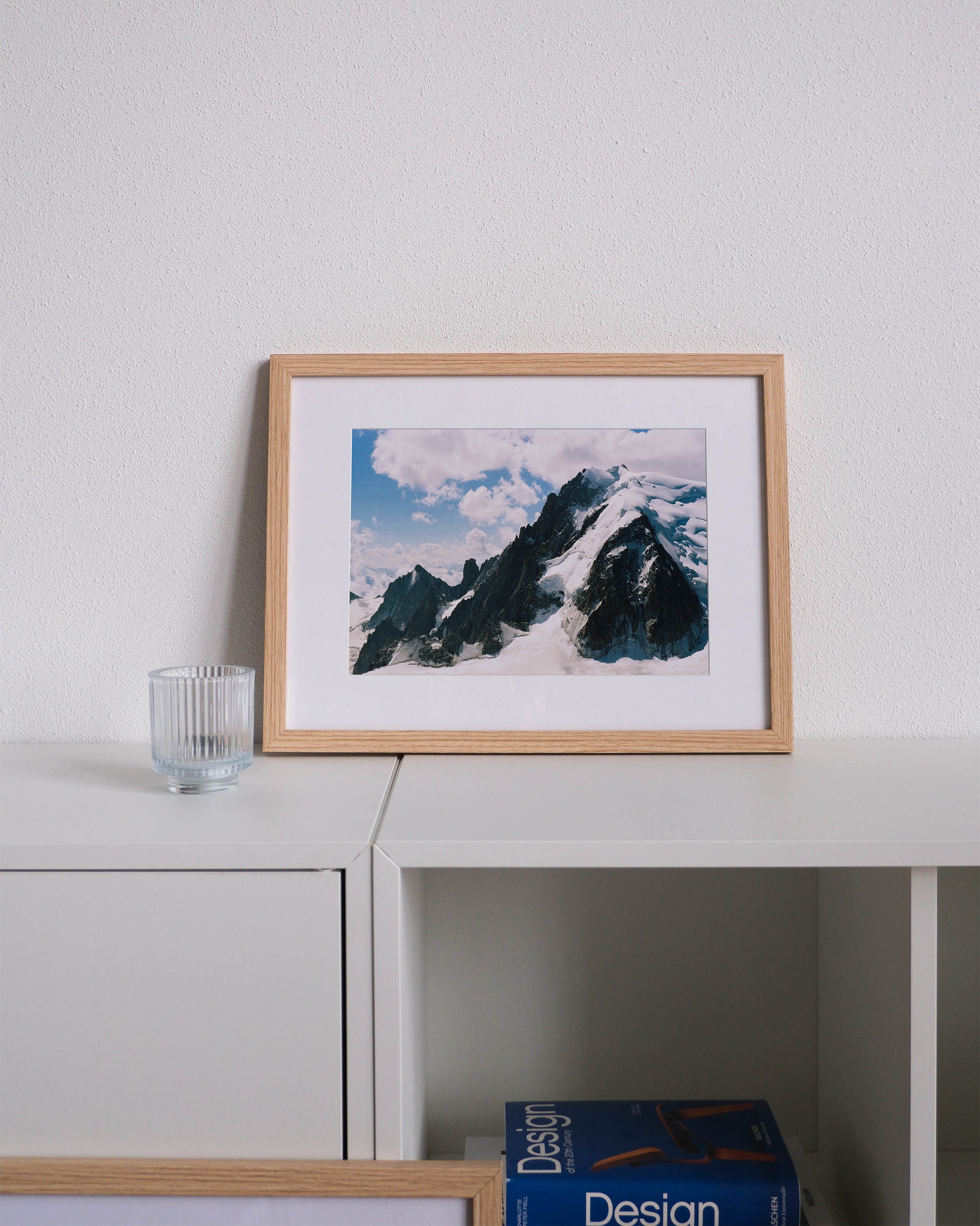 Framed analog photo print on Fujifilm paper featuring the towering peaks of the Mont Blanc massif, showcasing the rugged, snow-capped summits against the sky. Displayed on a modern white cabinet with a minimalist interior setting.