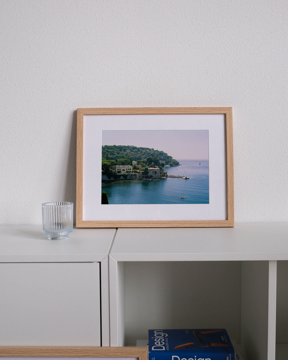 Framed analog photo print on Fujifilm paper featuring Saint-Jean-Cap-Ferrat on the French Riviera, with lush trees, paddle boarders on the Mediterranean, and luxurious villas along the coast, displayed on a modern white cabinet with a minimalist interior setting.