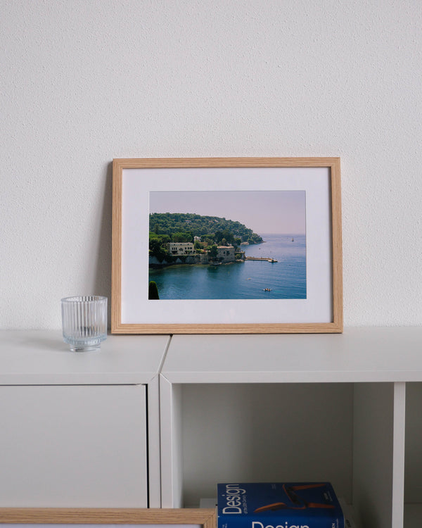 Framed analog photo print on Fujifilm paper featuring Saint-Jean-Cap-Ferrat on the French Riviera, with lush trees, paddle boarders on the Mediterranean, and luxurious villas along the coast, displayed on a modern white cabinet with a minimalist interior setting.