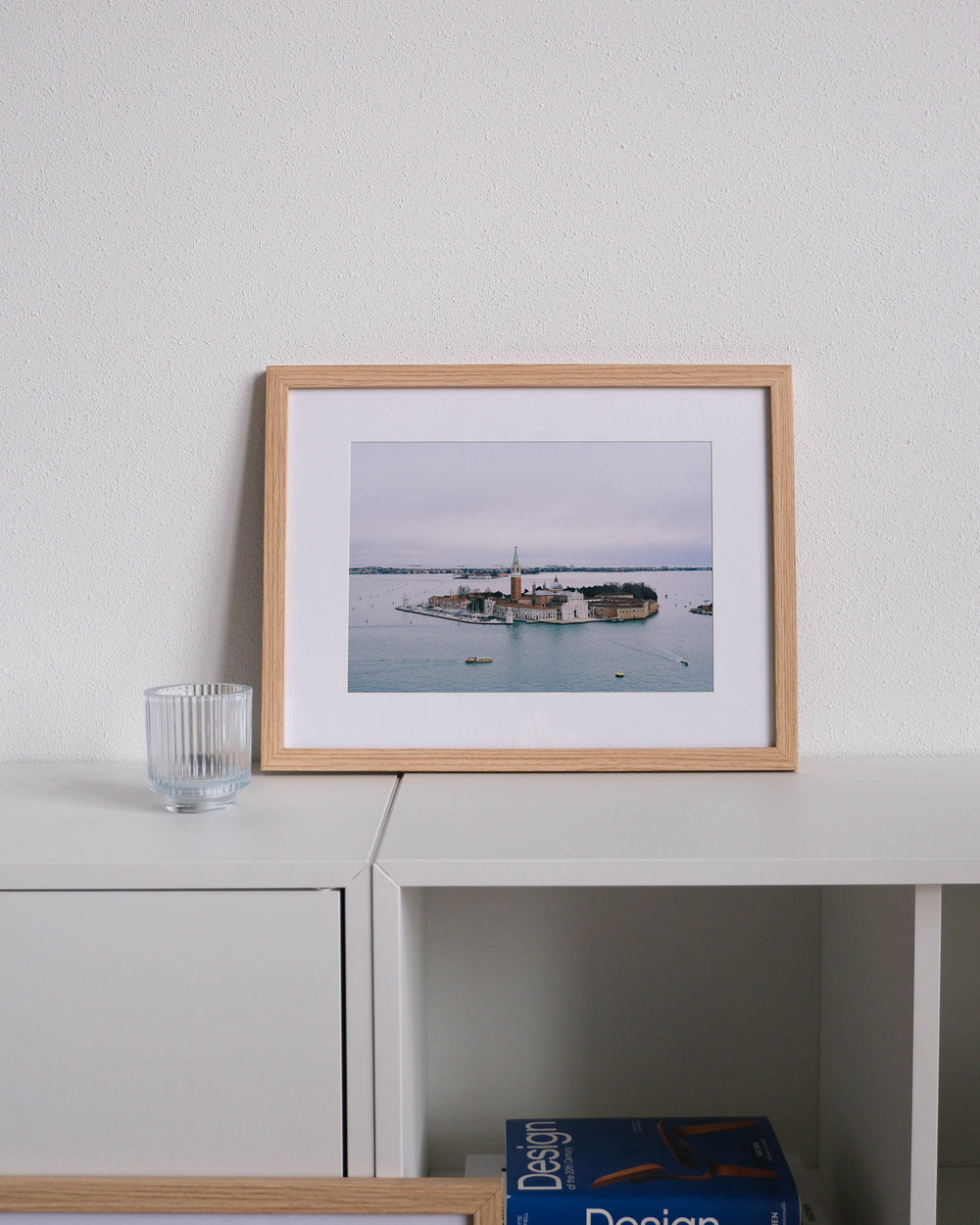 Framed analog photo print on Fujifilm paper featuring a view of Venice from the Loggetta del Sansovino, with the San Giorgio Maggiore Church standing out against the island. Boats float across the lagoon, and the cool winter light adds to the tranquil scene. Displayed on a modern white cabinet with a minimalist interior setting.