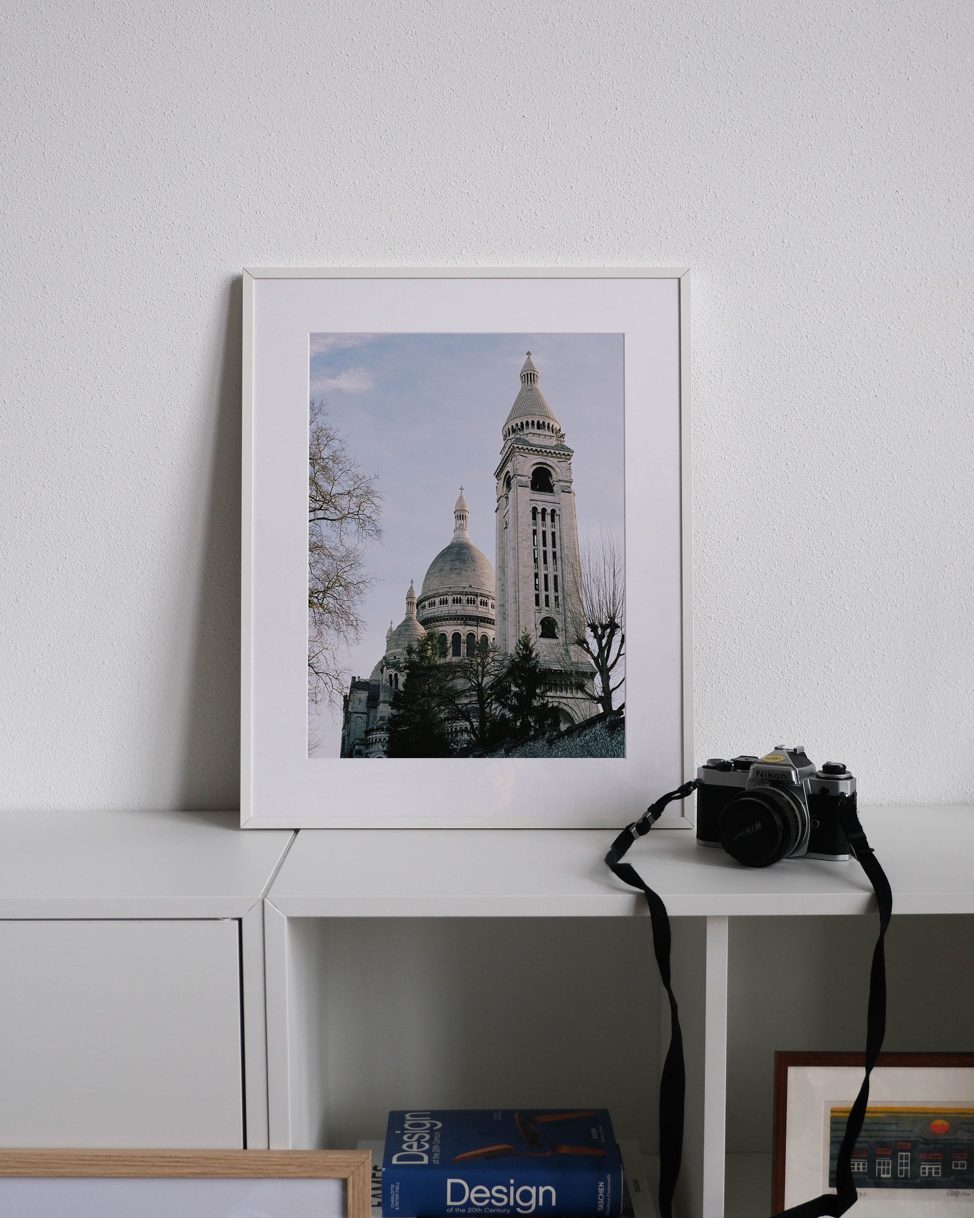 Framed analog photo print on Fujifilm paper featuring the towers of Sacré-Cœur in winter, bathed in sunlight with a clear blue sky above, displayed on a modern white cabinet with a minimalist interior setting.