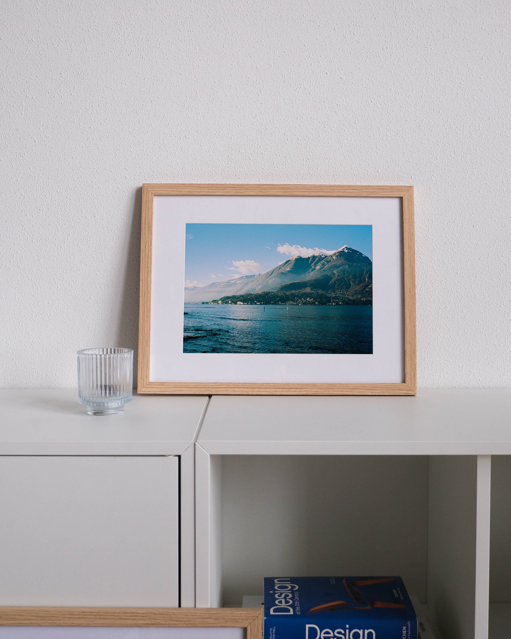 Framed analog photo print on Fujifilm paper featuring a mountain peak on Lake Como in winter, with mist drifting across the slopes and sunlight breaking through, displayed on a modern white cabinet with a minimalist interior setting.