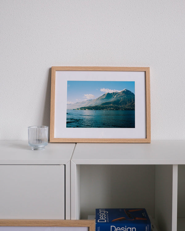 Framed analog photo print on Fujifilm paper featuring a mountain peak on Lake Como in winter, with mist drifting across the slopes and sunlight breaking through, displayed on a modern white cabinet with a minimalist interior setting.