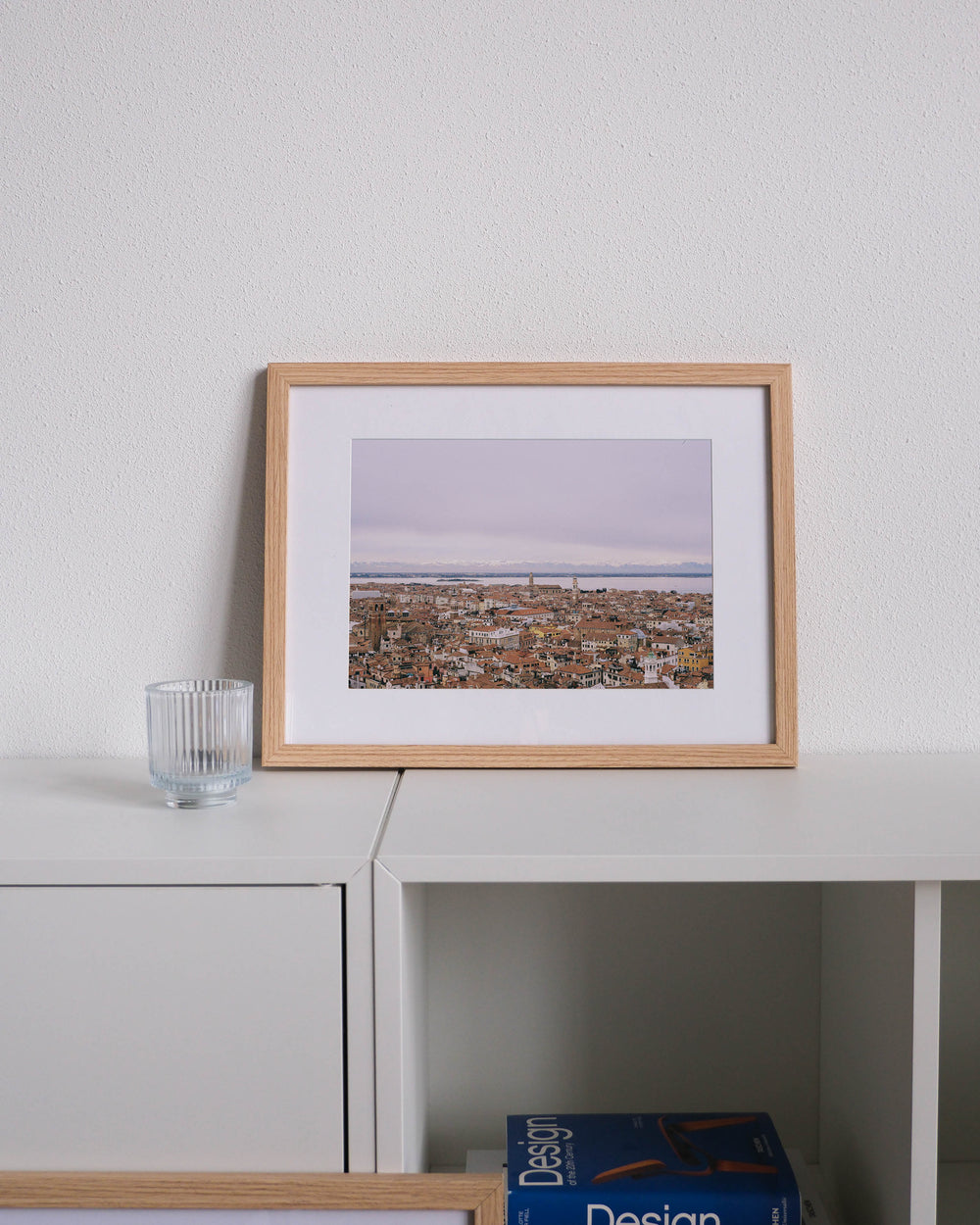 Framed analog photo print on Fujifilm paper featuring the iconic terracotta rooftops of Venice, captured from the tower of Loggetta del Sansovino. In the distance, the snow-capped peaks of the Alps rise above the lagoon, framed by a soft, clouded winter sky. Displayed on a modern white cabinet with a minimalist interior setting.