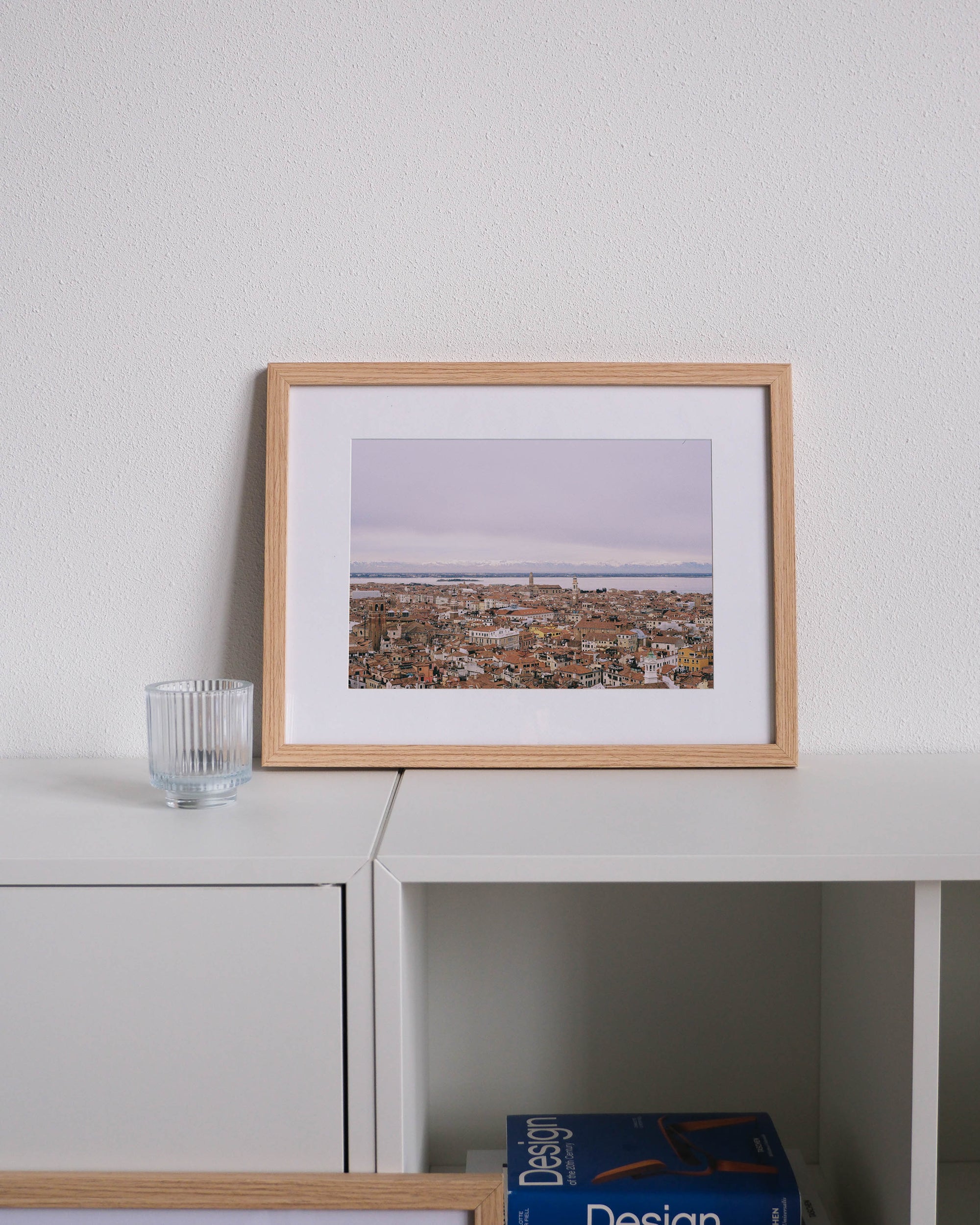 Framed analog photo print on Fujifilm paper featuring the iconic terracotta rooftops of Venice, captured from the tower of Loggetta del Sansovino. In the distance, the snow-capped peaks of the Alps rise above the lagoon, framed by a soft, clouded winter sky. Displayed on a modern white cabinet with a minimalist interior setting.