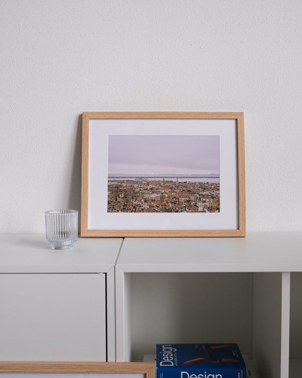 Framed analog photo print on Fujifilm paper featuring the iconic terracotta rooftops of Venice, captured from the tower of Loggetta del Sansovino. In the distance, the snow-capped peaks of the Alps rise above the lagoon, framed by a soft, clouded winter sky. Displayed on a modern white cabinet with a minimalist interior setting.