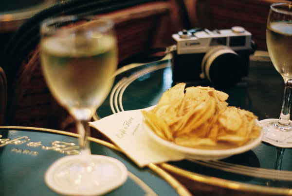Analog photo print featuring a close-up of an engraved table at Café du Flore, with a glass of white wine, a bowl of chips, and an analog camera, capturing a relaxed Parisian moment.