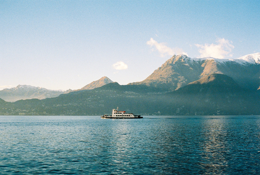 Analog photo print on Fujifilm paper featuring a ferry crossing Lake Como in winter in Bellano, with mountain peak Monte Bregagno in the background, partially covered in mist and touched by soft sunlight.