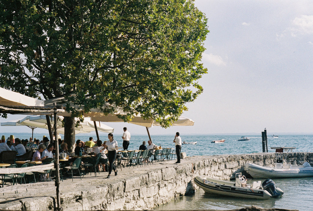 Analog photo print featuring a serene lakeside terrace on Lake Garda, with waiters serving guests under a tree, soft sunlight filtering through the leaves, and the tranquil waters in the background.