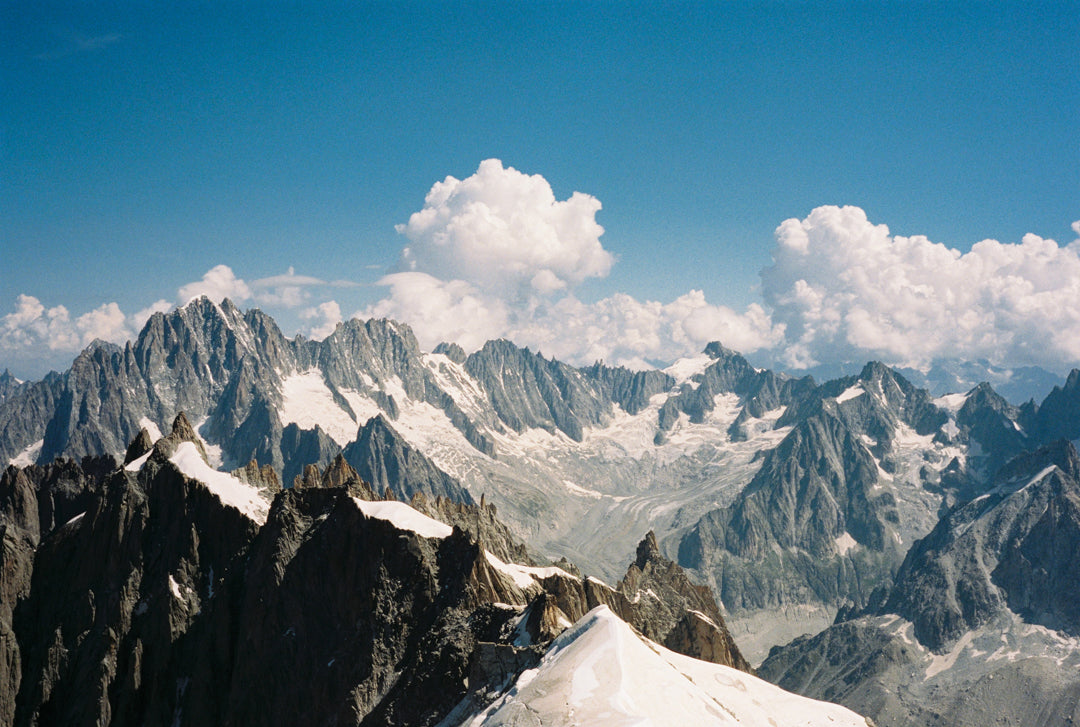 Analog photo print on Fujifilm paper featuring the Mont Blanc massif, captured from the Aiguille du Midi at 3842 meters. The steep peaks rise against a blue sky, with swirling clouds adding contrast to the rugged landscape. Captured in Chamonix.