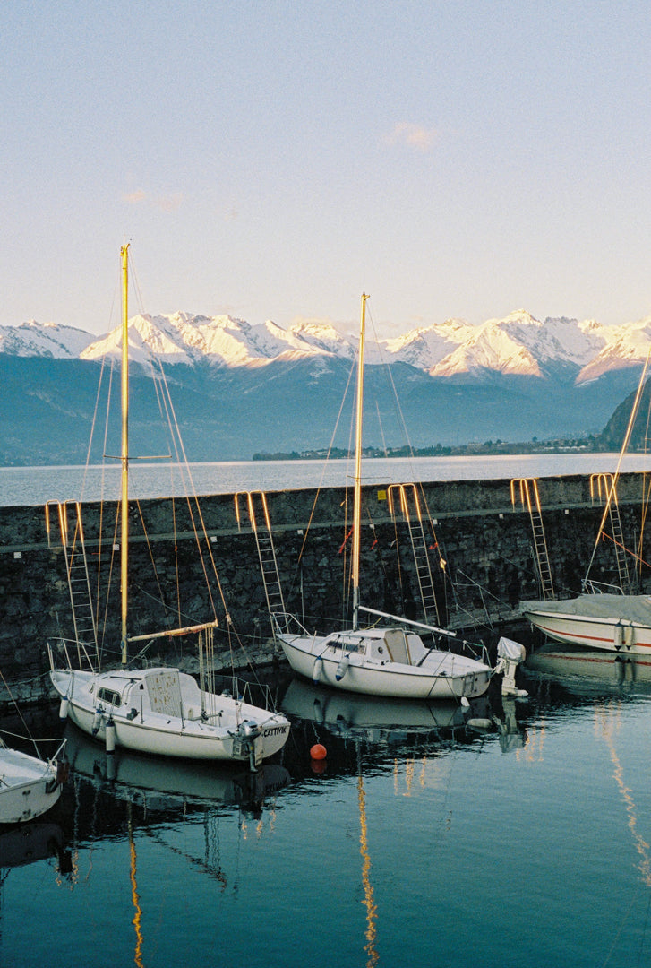 Analog photo print on Fujifilm paper featuring sailboats resting on the still water in Bellano Marina on a winter day, with mountains in the background and a warm sunset glow. 