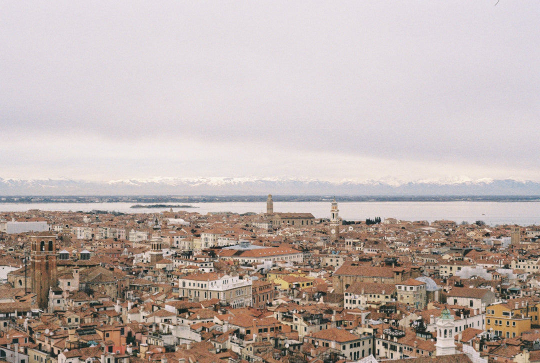 Analog photo print on Fujifilm paper featuring the iconic terracotta rooftops of Venice, captured from the tower of Loggetta del Sansovino. In the distance, the snow-capped peaks of the Alps rise above the lagoon, framed by a soft, clouded winter sky.